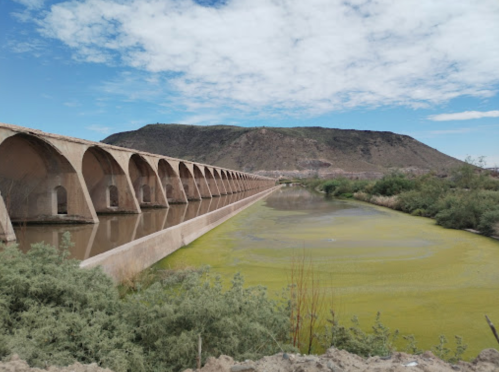 A long, arched bridge spans a river with green algae, set against a backdrop of a mountain and cloudy sky.